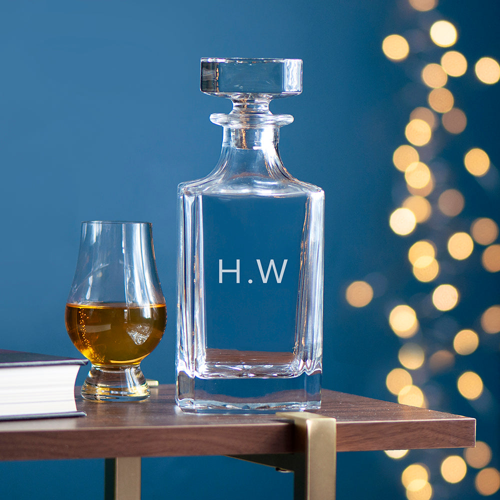 Clear glass decanter with 'H.W.' engraving next to a filled glass on a wooden surface with a blurred bokeh background.