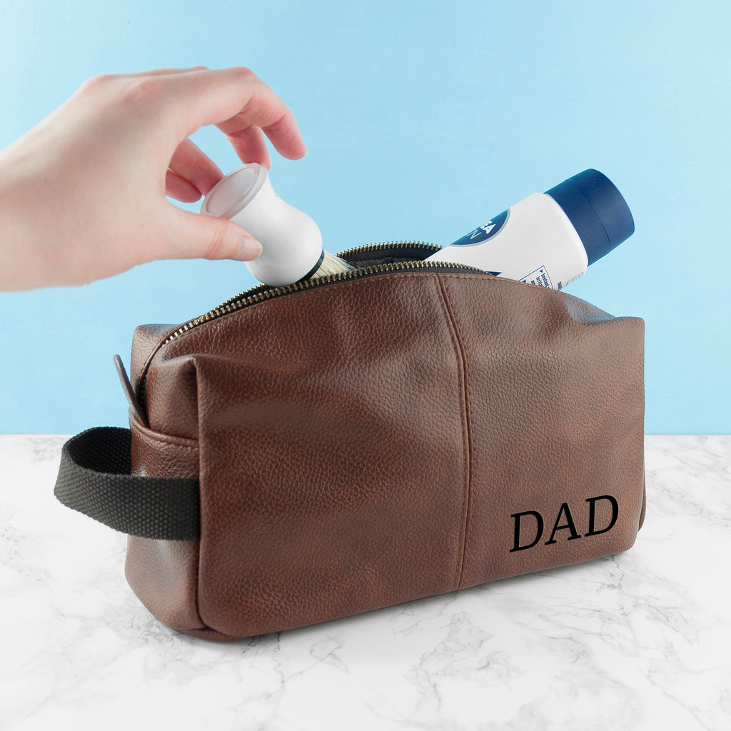 Brown leather toiletry bag with 'DAD' engraving on a marble surface against a light blue background.