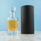 Clear glass decanter with amber liquid and 'Decant' label next to a black cylindrical container on a light blue background.