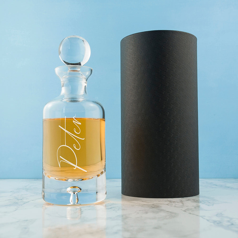 Clear glass decanter with amber liquid and 'Decant' label next to a black cylindrical container on a light blue background.