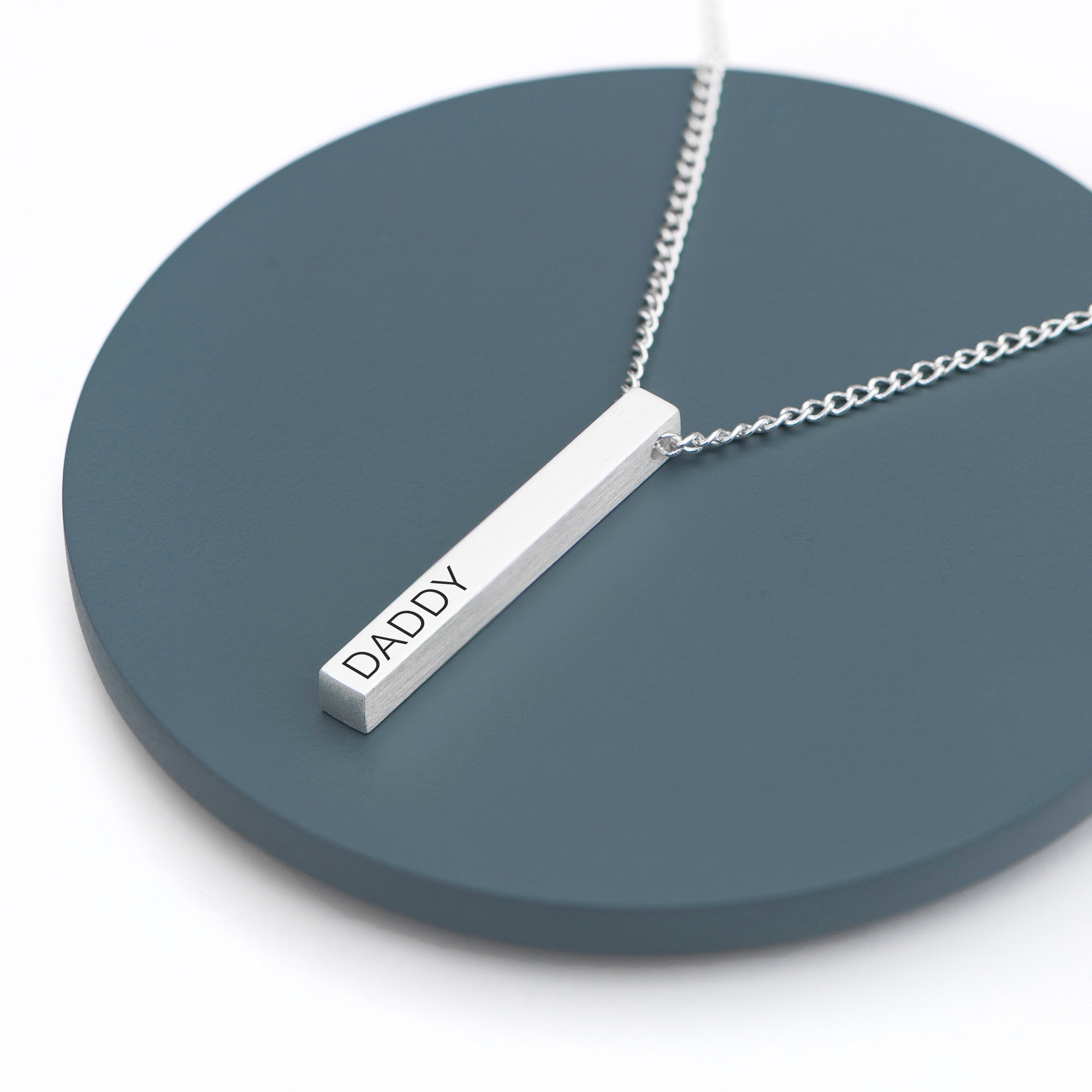 Silver necklace with a bar pendant inscribed 'DADDY' on a dark blue circular stand.