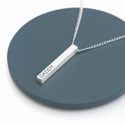 Silver necklace with a bar pendant inscribed 'DADDY' on a dark blue circular stand.