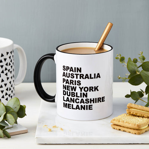White mug with black text and black handle, filled with coffee, on a white surface with cookies and plants.