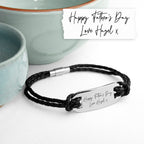 Black braided bracelet with engraved message on a white surface with ceramic bowls in the background.