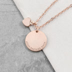 Rose gold necklace with a round pendant and heart charm on a gray and white background