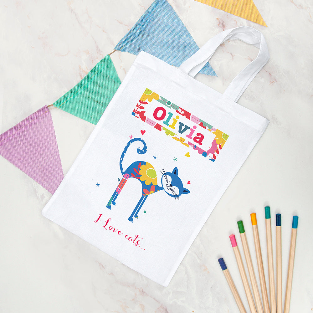 White tote bag with colorful cat design and text, placed on a textured surface with colored pencils.