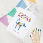 White tote bag with colorful cat design and text, placed on a textured surface with colored pencils.
