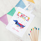 White tote bag with colorful dachshund design and text, placed on a textured surface with colored pencils.