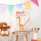 Children's drawstring bag with a giraffe design and 'Maisie' text, placed on a wooden stool with a teddy bear and books in the background.