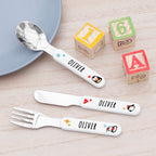 Children's cutlery set with 'OLIVER' on a wooden surface with blocks in the background