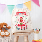 Drawstring bag with 'Scarlet' design, teddy bear, and stationery items on a wooden stool.