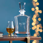 Clear glass decanter with 'H.W.' engraving next to a filled glass on a wooden surface with a blurred festive background.