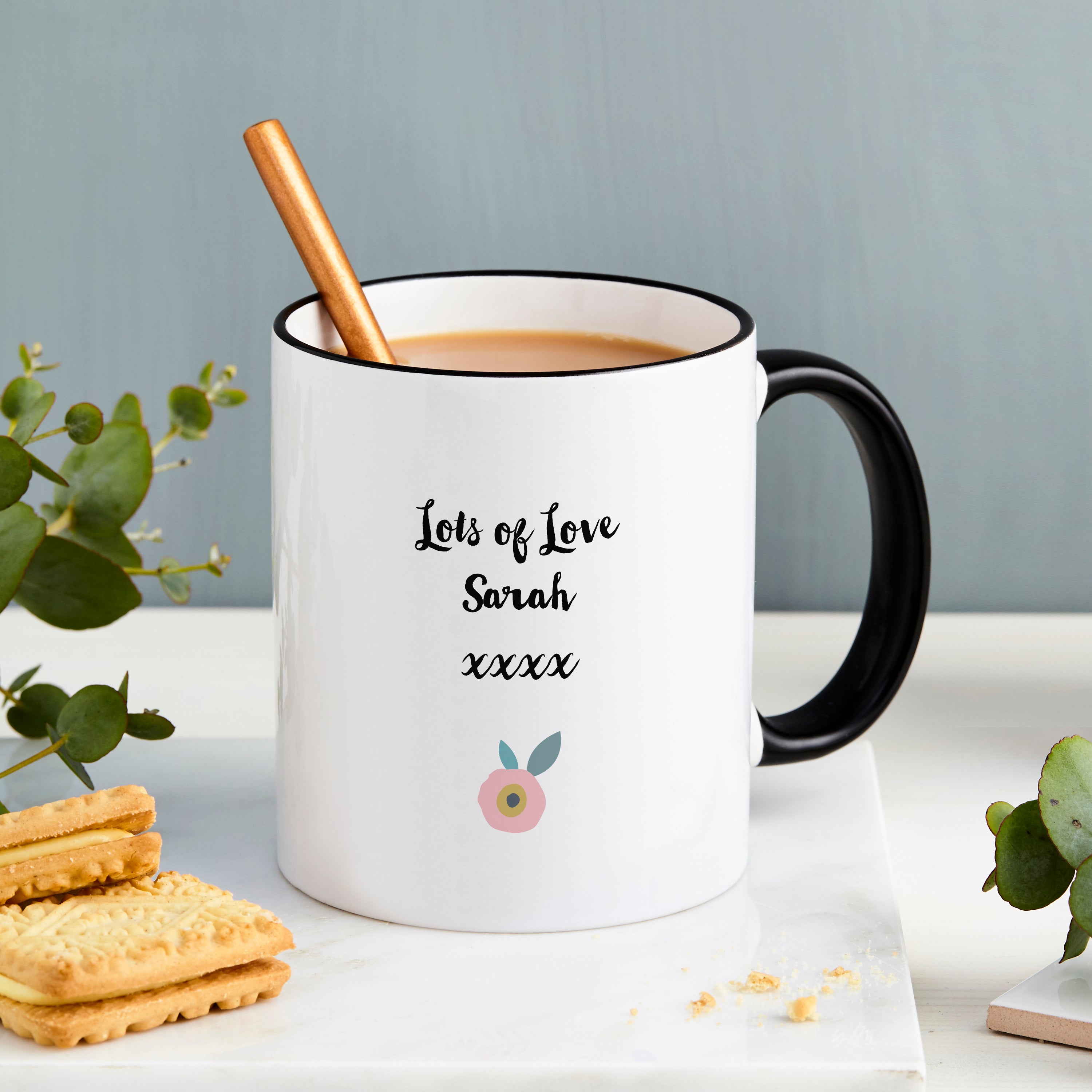 White mug with black handle and text, filled with a beverage, on a light surface with cookies and a plant.