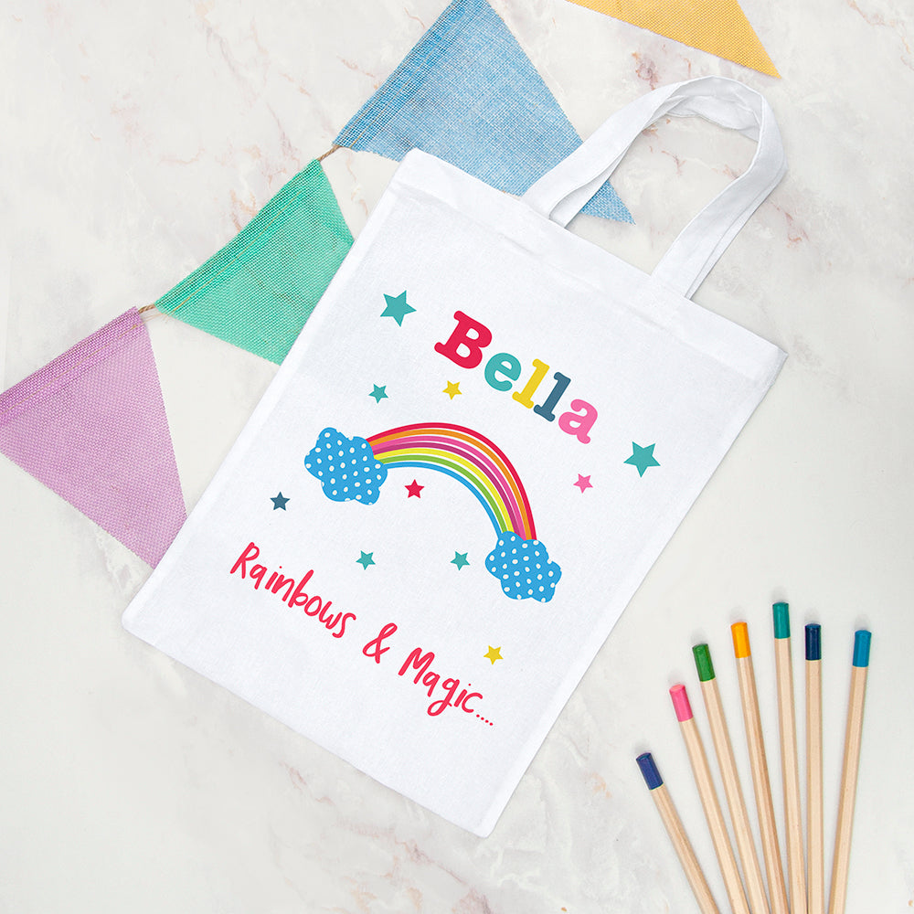 White tote bag with colorful rainbow design and text, surrounded by fabric swatches and colored pencils.