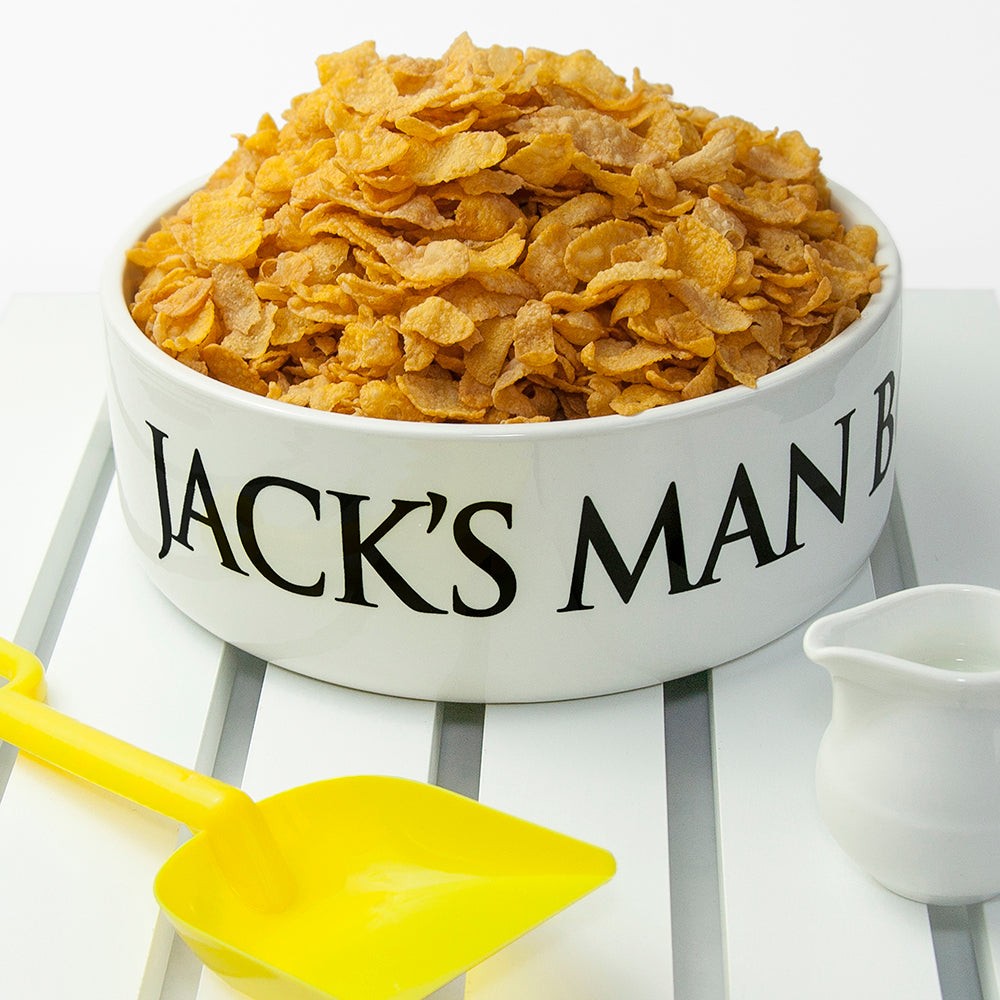 White bowl labeled 'Jack's Man Bowl' filled with cornflakes, accompanied by a yellow spoon and small white pitcher on a white background.