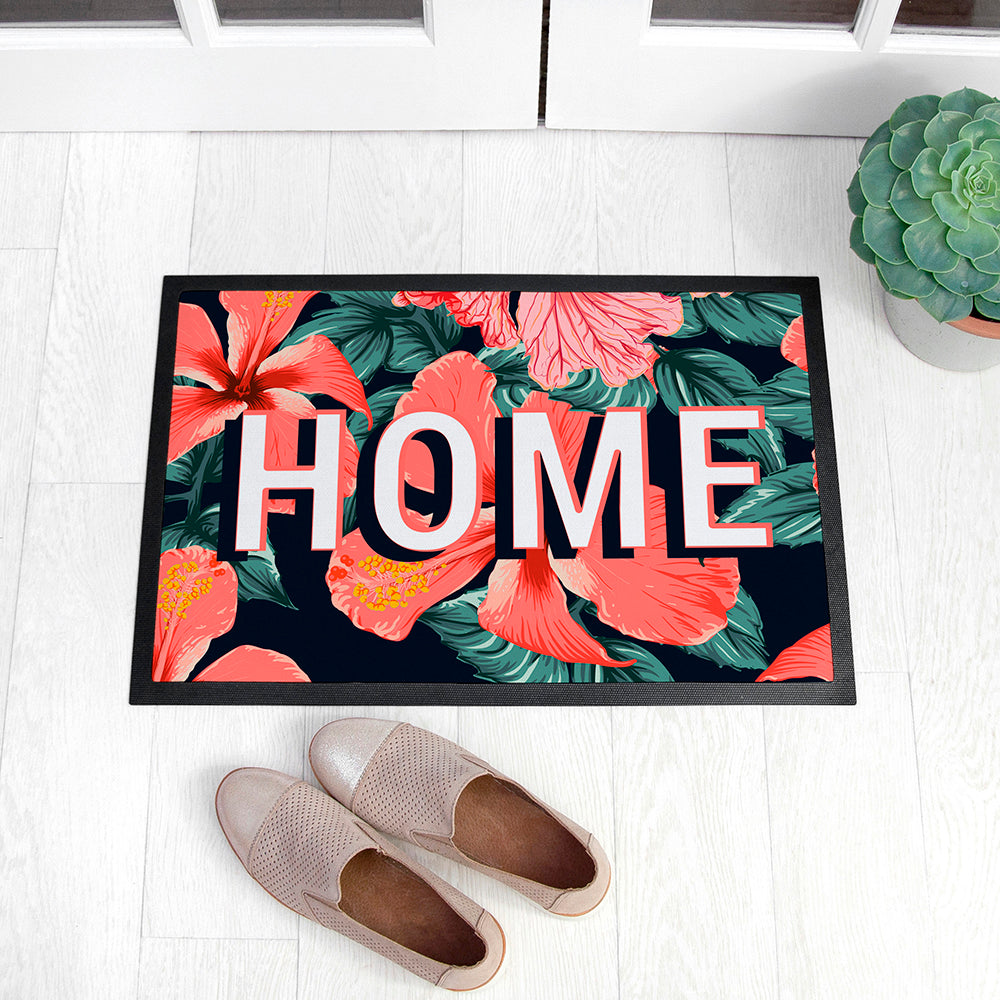 Decorative doormat with floral design and 'HOME' text on a white floor with shoes and a plant.