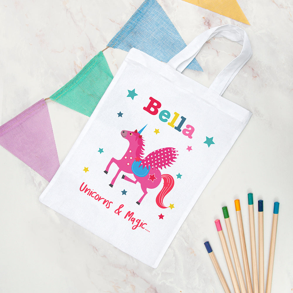 White tote bag with a pink unicorn design and 'Bella' text, surrounded by colorful pencils on a textured surface.
