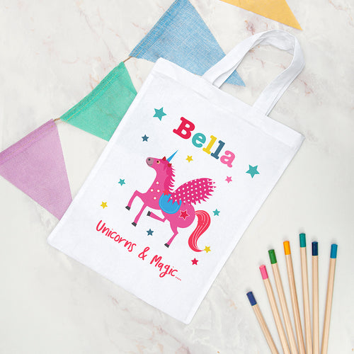 White tote bag with a pink unicorn design and 'Bella' text, surrounded by colorful pencils on a textured surface.