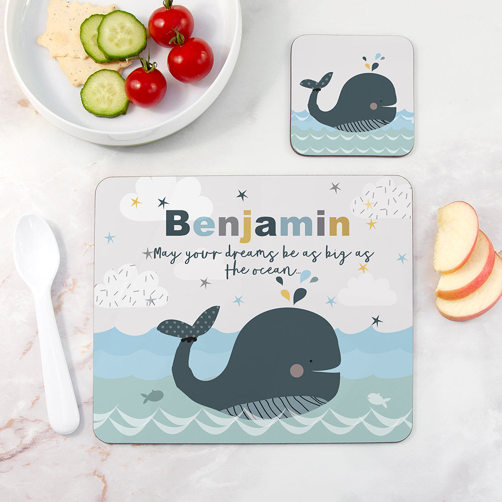 Placemat with whale design and 'Benjamin' text on a table with a bowl of fruit.