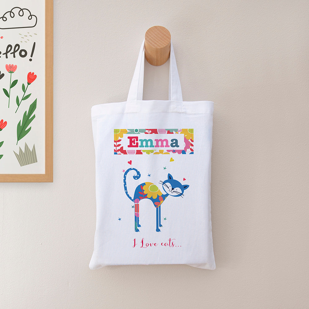 White tote bag with colorful cat design and 'Emma' text, hanging on a wooden hook.