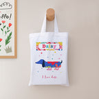 White tote bag with colorful dog illustration and text, hanging on a wooden hook against a light wall.