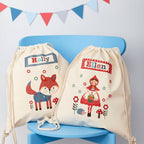 Two drawstring bags with embroidered designs of a fox and a fairy tale character on a blue chair.