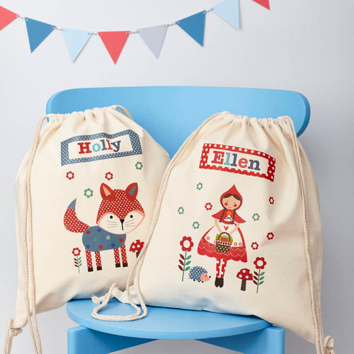Two drawstring bags with embroidered designs of a fox and a fairy tale character on a blue chair.