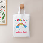 White tote bag with colorful design and text hanging on a wooden hook against a light wall.