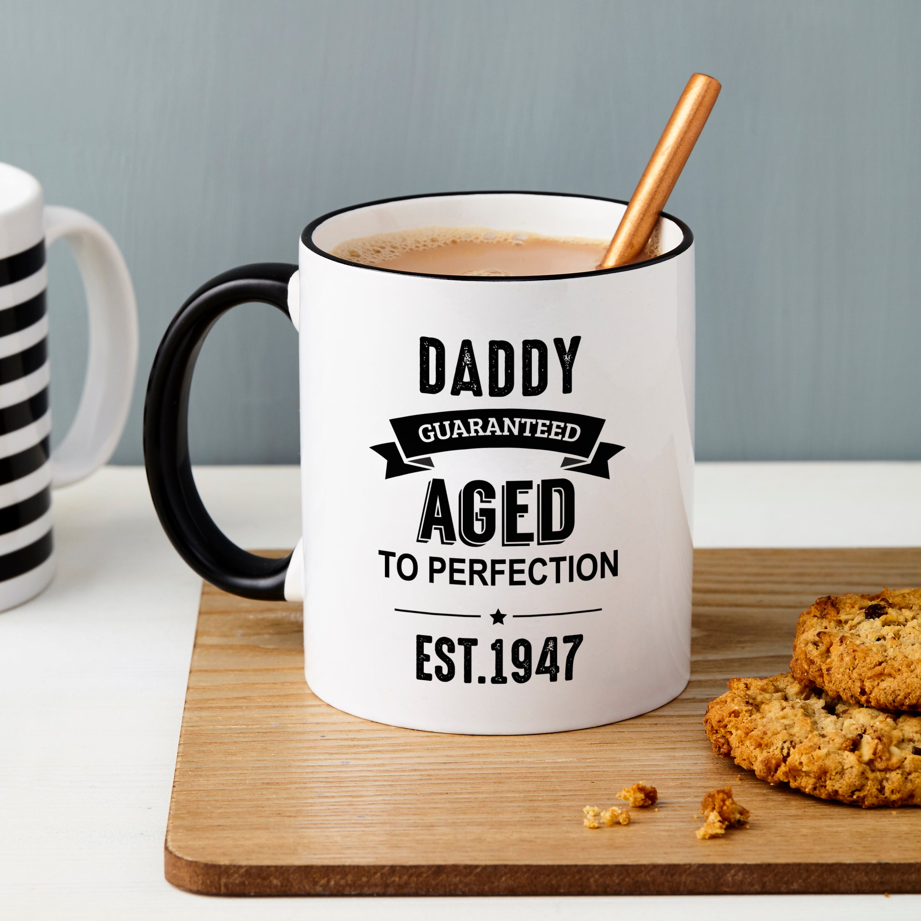 White mug with black text 'Daddy Guaranteed Aged to Perfection Est. 1947' on a wooden board with cookies.
