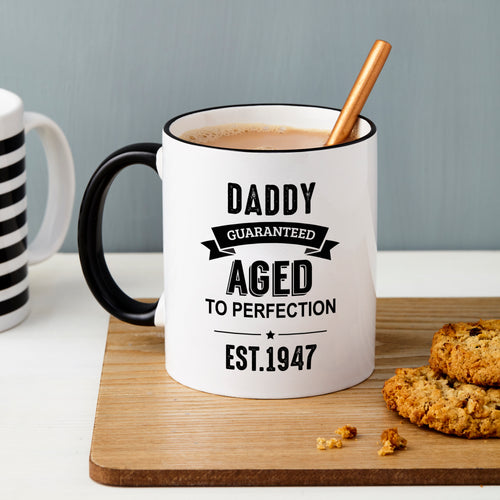 White mug with black text 'Daddy Guaranteed Aged to Perfection Est. 1947' on a wooden board with cookies.