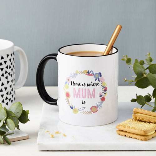 White mug with floral design and 'Home is where Mum is' text on a table with cookies and plants.